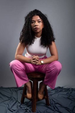 Güzel bir kadın, kıvırcık saçlı, gri arka planda oturmuş kameraya ciddi ciddi bakıyor..