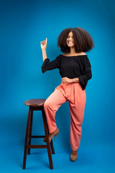 stock image Young woman with black power hair, leaning on a wooden stool pointing upwards. Isolated on blue background.