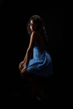 Düz saçlı, gözlüklü, mavi elbiseli fotoğraf için poz veren genç bir kadın. Siyah arkaplanda izole.