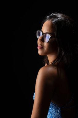 Düz saçlı, gözlüklü, mavi elbiseli fotoğraf için poz veren genç bir kadın. Siyah arkaplanda izole.