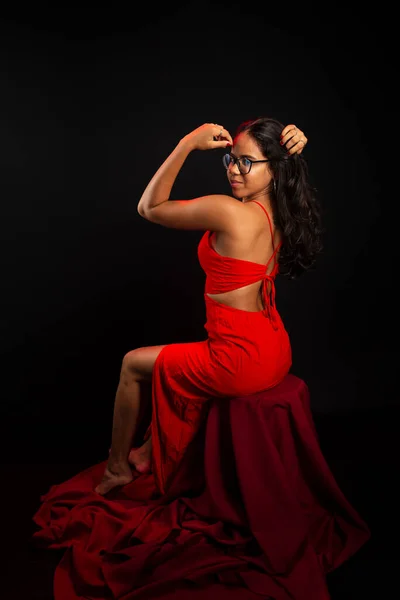 stock image Beautiful young woman dressed in red sitting with her hand in her hair, wearing prescription glasses and posing for a photo. Isolated on black background.