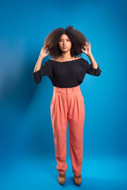 Güzel genç bir kadın, ayakta, günlük giysiler içinde, siyah güç saçlarına dokunuyor. Mavi arkaplanda izole.