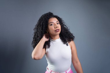 Genç ve güzel bir kadın kıvırcık saçlarına dokunuyor ve gri arka planda kameraya bakıyor..