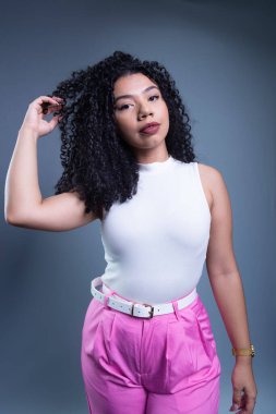 Genç ve güzel bir kadın kıvırcık saçlarına dokunuyor ve gri arka planda kameraya bakıyor..