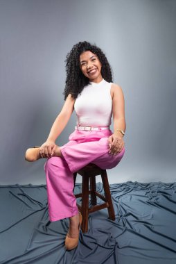 Kıvırcık saçlı, resmi giyinmiş güzel bir kadın tahta taburede oturmuş fotoğraf için poz veriyor. Gri arkaplanda izole.