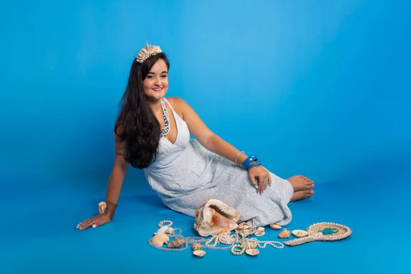 Portrait of beautiful black-haired woman wearing white clothes, sitting against blue background next to several sea shells and pearl necklaces. Devotee of iemanja.