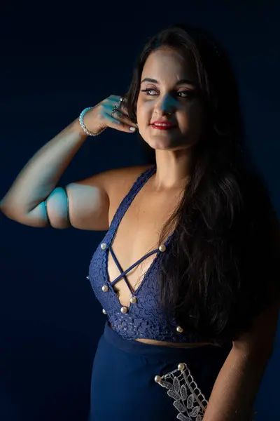 Portrait of a beautiful woman with black hair and blue clothes, with her hand close to her face posing for a photo. Isolated on dark blue background.