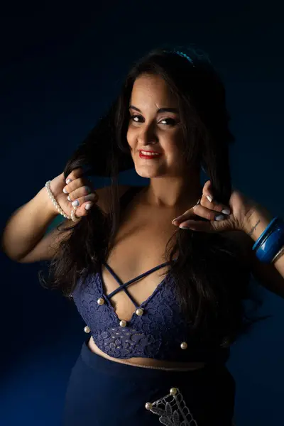 Portrait of a beautiful woman with black hair and blue clothes, with her hand close to her face posing for a photo. Isolated on dark blue background.