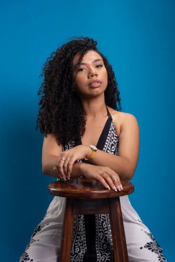 Tahta bir tabureye yaslanmış fotoğraf için poz veren kıvırcık saçlı güzel bir genç kadın. Mavi arkaplanda izole.