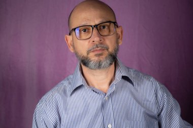 Kel, sakallı bir adam pembe arka planda kameraya bakıyor..