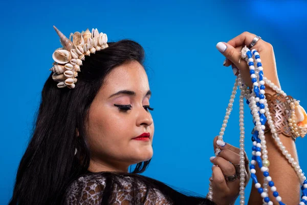 Beautiful, black-haired woman with shell tiara on her head holding and looking at pearl necklaces. Characterization of Iemanja.