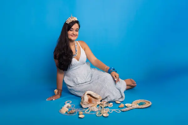 Portrait of beautiful black-haired woman wearing white clothes, sitting against blue background next to several sea shells and pearl necklaces. Devotee of iemanja.
