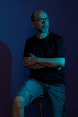 Sakallı, gözlüklü kel bir adamın portresi. Tahta taburede oturmuş fotoğraf için poz veriyor. Koyu leylak arka planda izole edilmiş.