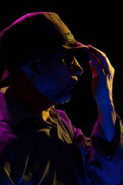 Şapka takan, güneş gözlüğü takan, sakallı ve elleriyle jest yapan gizemli bir adamın portresi. Stüdyoda renkli ışıklar ve koyu arkaplan ile portre.