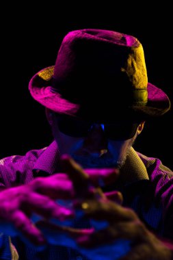 Şapka takan, güneş gözlüğü takan, sakallı ve elleriyle jest yapan gizemli bir adamın portresi. Stüdyoda renkli ışıklar ve koyu arkaplan ile portre.