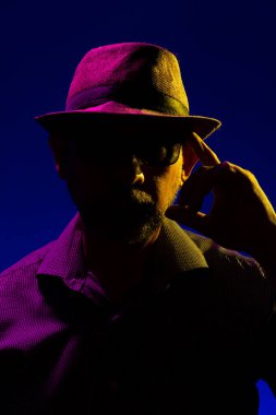Yüzüne dokunan şapka ve güneş gözlüğü takan ciddi ve gizemli bir adamın portresi. Stüdyoda renkli ışıklar ve koyu arkaplan ile portre.