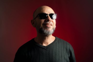 Beyaz, kel, güneş gözlüklü, kendine güvenen, gülümseyen bir adam. Kırmızı arkaplanda izole.