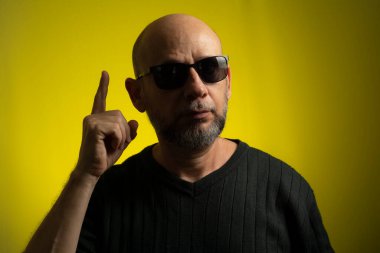 Kel, sakallı, güneş gözlüğü takan ve elleriyle işaret yapan olgun bir adamın portresi. Sarı arkaplanda izole edilmiş.