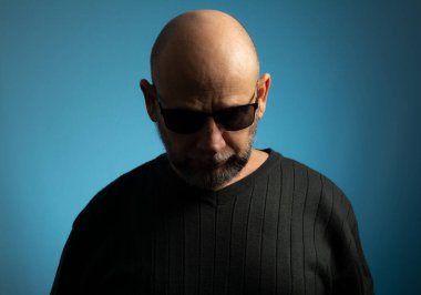 Güneş gözlüklü kel ve sakallı olgun bir adamın portresi aşağıya bakıyor. Mavi arkaplanda izole.