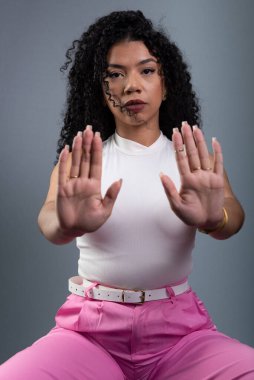 Güzel bir kadın oturuyor, iki eliyle dur işareti yapıyor. Gri arkaplandaki kameraya bakıyorum..