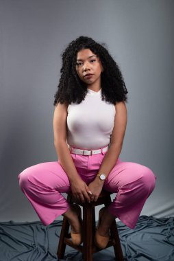 Güzel bir kadın, kıvırcık saçlı, gri arka planda oturmuş kameraya ciddi ciddi bakıyor..