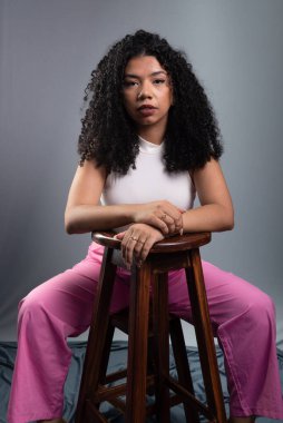 Güzel bir kadın, kıvırcık saçlı, gri arka planda oturmuş kameraya ciddi ciddi bakıyor..