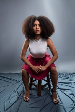 Güzel bir genç kadın, siyah saçlı, tahta bir taburede oturmuş, kameraya ciddi ciddi bakıyor. Gri arkaplanda izole.