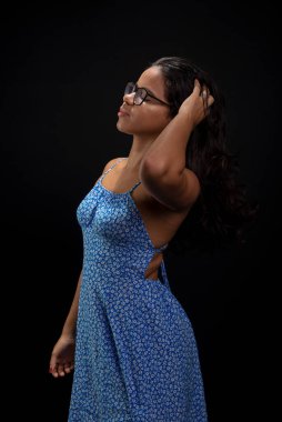 Gözlüklü güzel esmer kadın saçlarına dokunup poz veriyor. Siyah arka planda mavi elbiseler giyiyor..