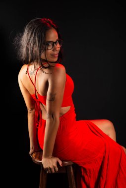Gözlüklü, kırmızı elbiseli, fotoğraf için poz veren güzel bir genç kadın. Siyah arkaplanda izole.