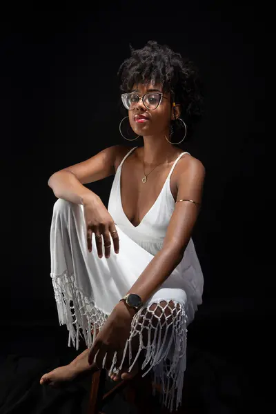 Beautiful woman, with black hair, glasses, sitting wearing white clothes and posing for the camera. isolated on black background.