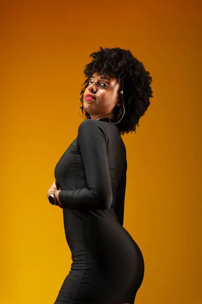 Young, sensual woman, with black power hair, standing, wearing black clothing and posing for a photo. Isolated on yellow background.