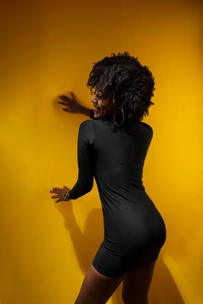 Young sensual woman with black power hair, wearing black clothes leaning against a yellow wall posing for a photo.