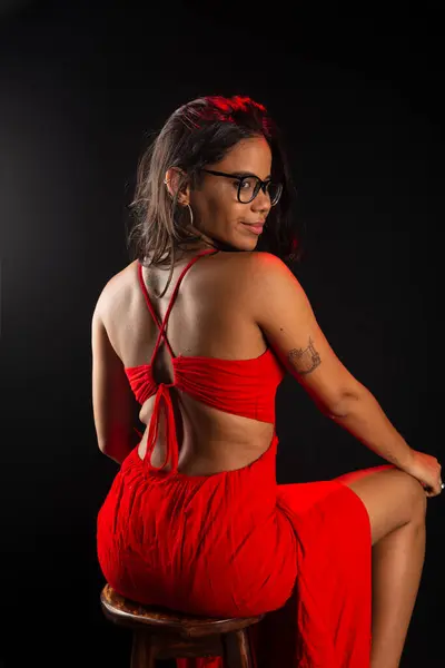 Beautiful young woman wearing glasses wearing red color outfit sitting posing for photo. Isolated on black background.