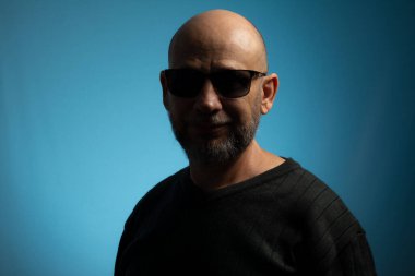 Kel ve sakallı yetişkin bir adamın portresi. Güneş gözlüğü takmış kameraya bakıyor. Mavi arkaplanda izole.