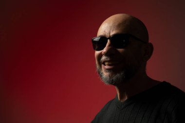 Yakışıklı, kel, sakallı, güneş gözlüğü takan, gülümseyen ve kendinden emin bir adam. Kırmızı arkaplanda izole.