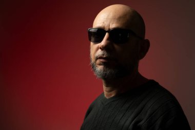 Yakışıklı, kel ve sakallı, güneş gözlüğü takan ciddi ve kendine güvenen bir adam kameraya bakıyor. Kırmızı arkaplanda izole.