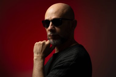 Güneş gözlüklü, yakışıklı, kel ve sakallı bir adam. Ciddi ve kendine güvenen bir poz veriyor. Kırmızı arkaplanda izole.
