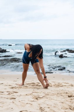 Güzel, kıvırcık saçlı, spor kıyafetleriyle kumsalda esneme hareketleri yapan genç bir kadın. Sağlıklı yaşam tarzı.
