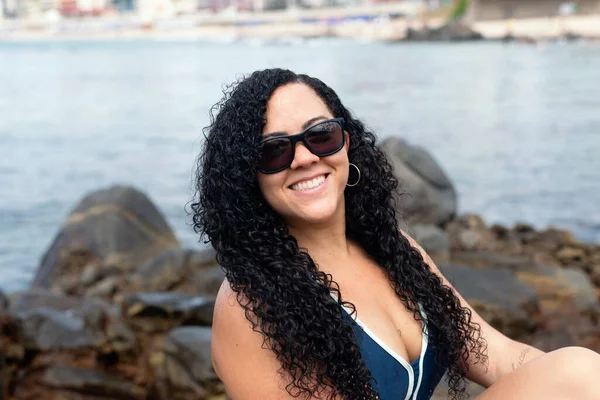 stock image Beautiful, curly-haired woman wearing sunglasses looking at the camera against the sea in the background. Happy person traveling.