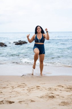 Fitness kadını, ayakta duruyor, bacak egzersizleri yapıyor kumsaldaki kumlara bakan kameraya bakıyor ve bulutlu gökyüzüne karşı. Sağlıklı yaşam tarzı.