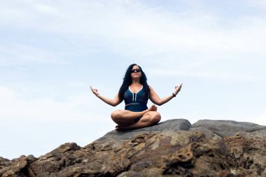 Güzel, kıvırcık saçlı bir kadın plajda oturmuş yoga egzersizi yapıyor. Seyahat eden mutlu kişi.