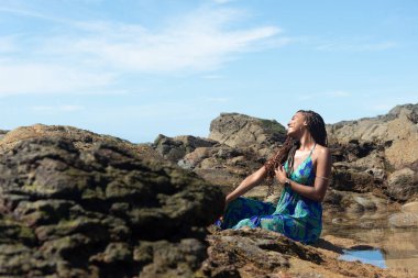 Güzel bir kadın, mavi elbiseli, elleri örgülü kayalıklarda oturuyor, kumsalda güneşleniyor ve yan tarafa bakıyor. Mavi gökyüzüne karşı.