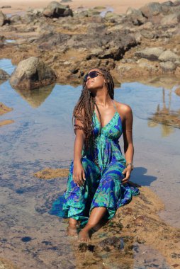 Güzel bir kadın, saç örgüleri ve mavi elbisesiyle kumsalda güneşlenen kayalarda oturuyor. Seyahat eden kişi.