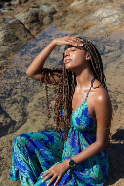 Mavi elbiseli güzel bir kadın kayalıklarda oturuyor, gözleri kapalı saçlarına dokunuyor, kumsalda güneşleniyor. Mavi gökyüzüne karşı.