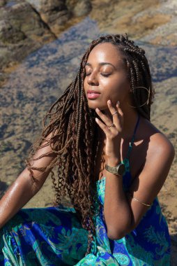 Güzel bir kadının portresi, örgülü ve mavi elbiseli, kumsalın kayalıklarında mavi gökyüzüne karşı poz veriyor..