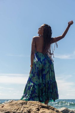 Güzel bir kadın, saç örgüleri ve mavi elbiseleriyle sahildeki kayalıklarda dikilip mavi gökyüzüne karşı poz veriyor..