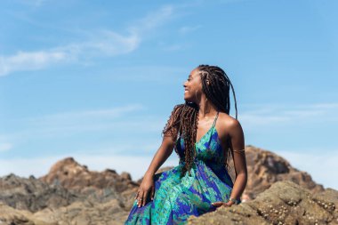 Güzel bir kadın, saç örgüleri ve mavi elbiseleriyle kumsalda güneşlenen kayalarda oturuyor ve mavi gökyüzüne karşı poz veriyor..