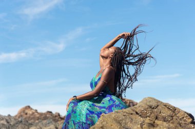 Mavi elbiseli güzel bir kadın kumsalda kayalıklarda güneşleniyor ve mavi gökyüzüne karşı örgülerine dokunuyor..