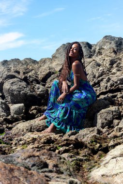 Mavi elbiseli, örgülü saçlı, kumsalın karanlık kayalarında oturmuş mavi gökyüzüne karşı güneşlenen güzel bir kadın..