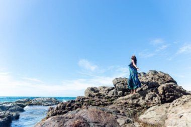 Güzel bir kadın, mavi elbiseli ve örgülü saçlı, kumsalın karanlık kayalıklarında duruyor, açık kollarıyla mavi gökyüzüne karşı..